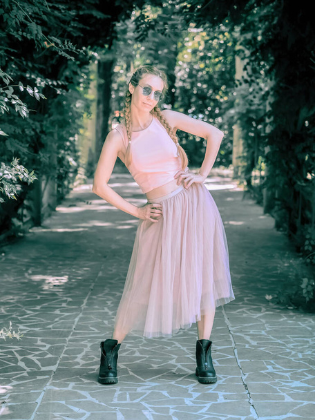 A lady with pigtails in a tutu and rough boots stands with her hands on her belt and legs apart in an alley in a summer park. Post-processing. Artistic effect. - Foto, immagini