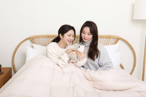 winter season concept, two beautiful Asian Korean women looking at their smartphones on bed - Photo, Image