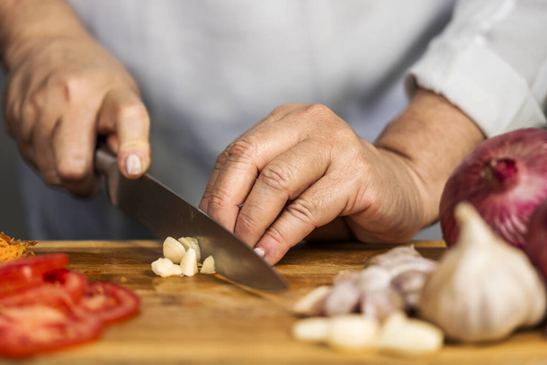 Close up view of the hands of an adult woman chopping a garlic with a kitchen knife on a cutting board for lasagna preparation. - Photo, image
