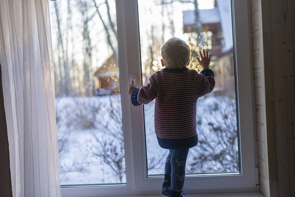 little boy stand near the window and look at the snowy street in winter, when it is very cold. - Фото, изображение