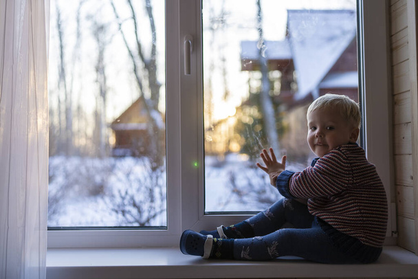 little boy stand near the window and look at the snowy street in winter, when it is very cold - Foto, afbeelding