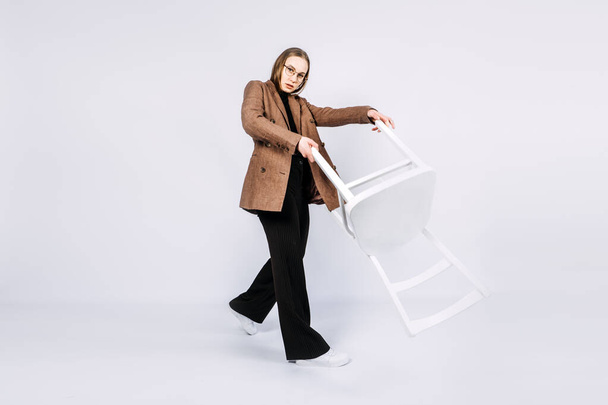 Full length studio portrait of exciting young woman fashion designer, stylist with white chair. Fashionable stylish blonde long hair girl in casual style cloth playing with chair. - Zdjęcie, obraz