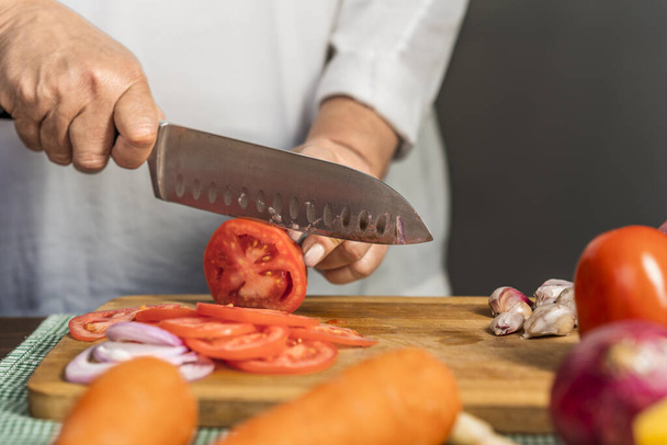 Close up view of the hands of an adult woman cutting tomatoes into slices with a kitchen knife on a wooden cutting board for lasagna preparation. - Foto, imagen