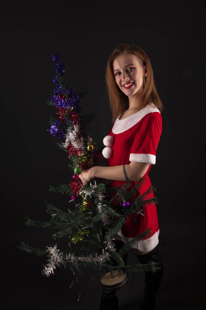 Vrolijk meisje in rode kerstman kostuum poseren met versierde kerstboom in studio - Foto, afbeelding