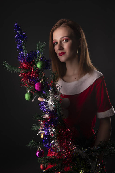 Vrolijk meisje in rode kerstman kostuum poseren met versierde kerstboom in studio - Foto, afbeelding