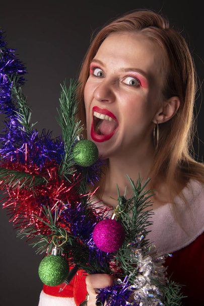 Cheerful girl in red Christmas Santa costume posing with decorated Christmas tree in studio - 写真・画像