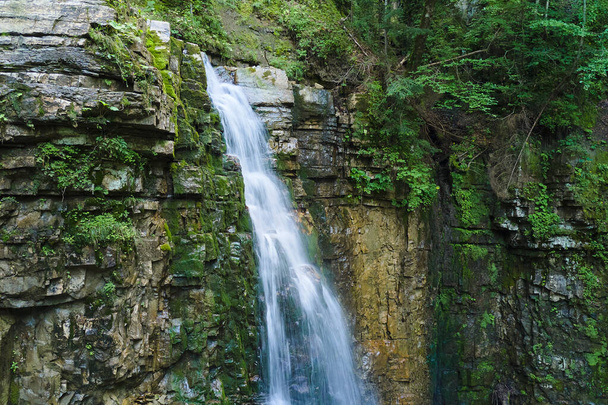 Водопад на горной реке с белой пенной водой, падающей с скалистого образования в летнем лесу - Фото, изображение