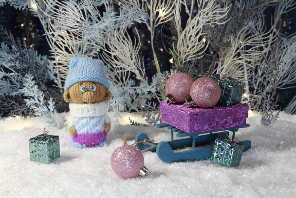 Un gnomo de juguete de punto se encuentra en la nieve artificial y un trineo de juguete con regalos se encuentra junto a él en el contexto de los árboles artificiales blancos y azules. Primer plano. Estudio nocturno rodaje, - Foto, imagen