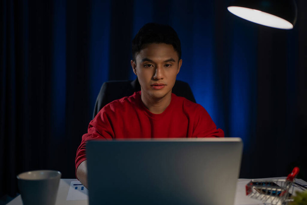 Handsome Asian man working on desk table looking at laptop computer in dark late night working feeling serious thinking and determinated at home office concept. - Foto, immagini