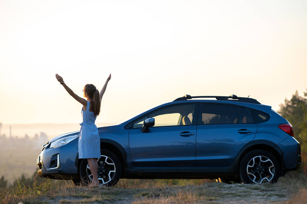 Gelukkige jonge vrouw bestuurder in blauwe jurk genieten van warme zomeravond staan naast haar auto. Reizen en vakantie concept - Foto, afbeelding