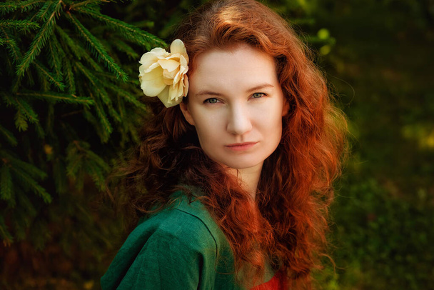 Portrait of a red-haired woman with a rose flower in her hair in the park. A curly-haired woman near the green fir branches. - Фото, изображение