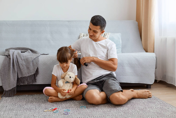 Indoor shot man with dark hair wearing white t shirt combing daughter's hair while sitting on floor near cough, preparing for going to school, expressing positive emotions, kid holding toy. - Foto, Imagen