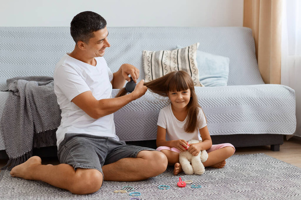 Indoor shot of attractive man wearing white t shirt sitting on floor and making hairstyle for his cute little daughter, taking care of his female child, spending pleasant time together. - Zdjęcie, obraz