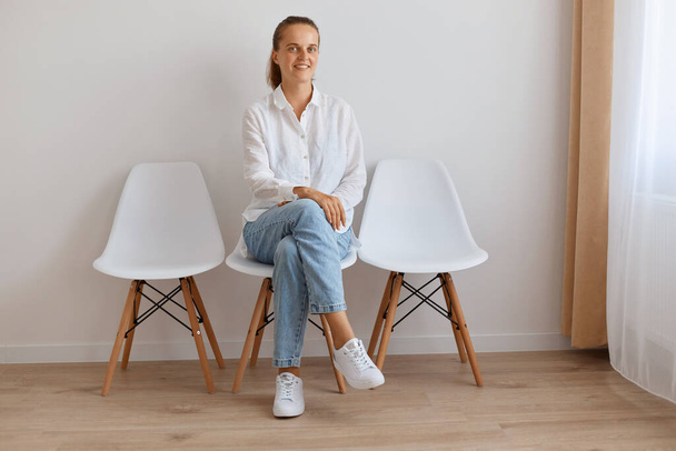 Image confident Caucasian young adult woman with ponytail wearing white shirt and jeans, sitting on chair against light wall indoor, posing with kind look and charming smile, expressing confidence. - Фото, изображение