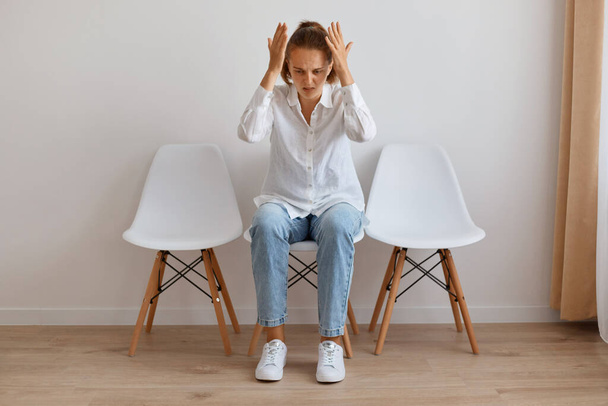 Horizontal shot of angry Caucasian young adult woman with ponytail wearing white shirt and jeans, sitting on chair against light wall indoor, raised hands, having problems, looks frustrated. - Foto, Imagem