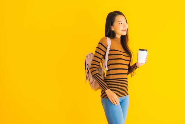 Portret mooie jonge aziatische vrouw met bagpack en koffiebeker in haar hand op geel geïsoleerde achtergrond - Foto, afbeelding