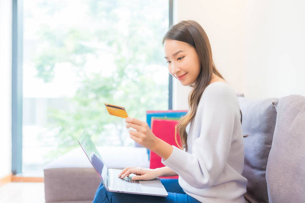 ポートレート美しい若いアジアの女性は自宅でオンラインショッピングのためのラップトップクレジットカードを使用します - 写真・画像