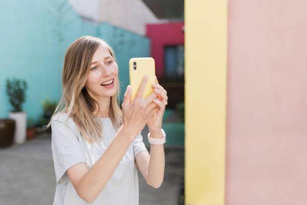 Pretty laughing caucasian woman with smartphone has good time outdoor. portrait of trendy millennial girl with blonde hair wears fashionable clothing - Photo, Image