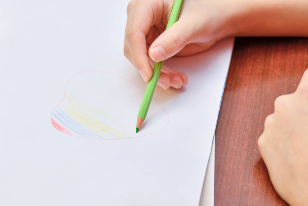 La ragazza disegna un cuore su un foglio bianco di carta e lo dipinge sotto forma di arcobaleno con matite colorate. - Foto, immagini