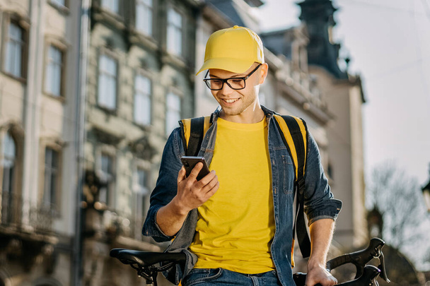 自転車に乗ってスマホでテキストメッセージを送る幸せなデリバリーマンのポートレート。彼は肩に黄色いリュックを担いで配達している。笑ってる. - 写真・画像