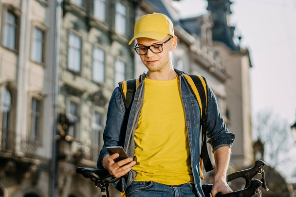 Portrait of a happy delivery man who is sitting on a bicycle and texting on the smartphone. He is carrying a yellow backpack for delivery on his shoulders. - Valokuva, kuva