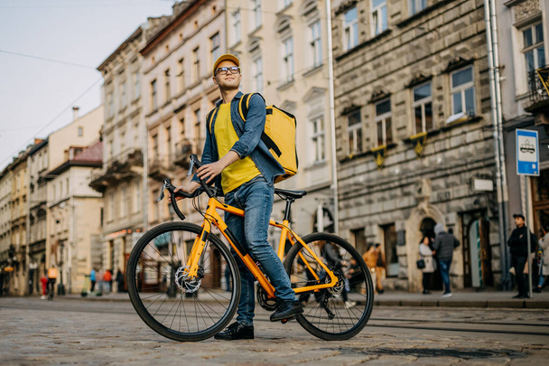Un repartidor feliz que está sentado en una bicicleta y mirando alrededor. Lleva una mochila amarilla en los hombros.. - Foto, imagen