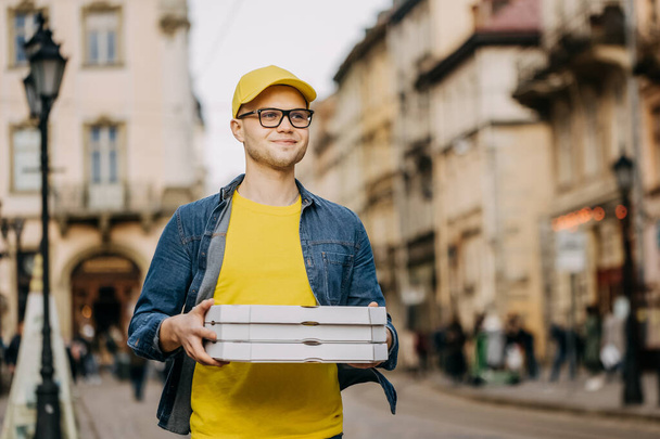 Молодий щасливий чоловік робить доставку трьох піц. Він у жовтій шапці та окулярах. Він проходить через центр старого міста
. - Фото, зображення