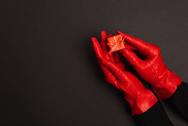 cropped view of woman in red gloves holding tiny gift box on black  - 写真・画像