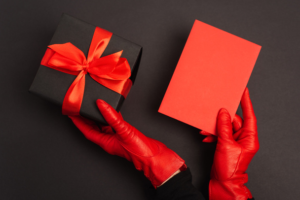 περικοπή άποψη του προσώπου σε κόκκινα γάντια που κατέχουν τυλιγμένο κουτί δώρου και το φάκελο σε μαύρο  - Φωτογραφία, εικόνα