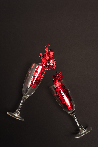 top view of champagne glasses with shiny confetti hearts on black  - Foto, Imagem