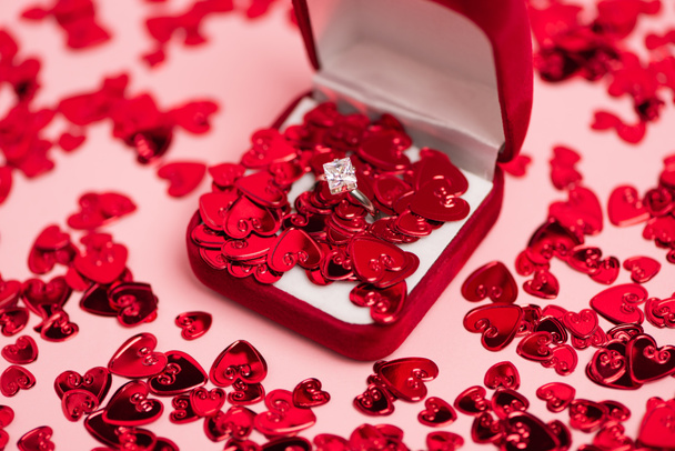close up of jewelry box with diamond ring near red confetti hearts on pink - Zdjęcie, obraz
