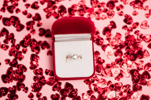 Вид сверху шкатулки с бриллиантовым кольцом рядом с блестящими сердечками на розовом фоне - Фото, изображение