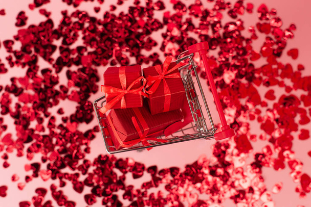 top view of small shopping cart with presents near shiny red confetti hearts on pink - Фото, изображение