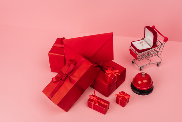 high angle view of red wrapped gift boxes and envelope near shopping cart with engagement ring on pink  - Fotoğraf, Görsel