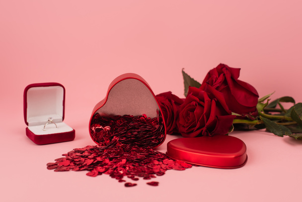 boîte métallique en forme de coeur avec confettis près de roses rouges et bague de fiançailles sur rose  - Photo, image