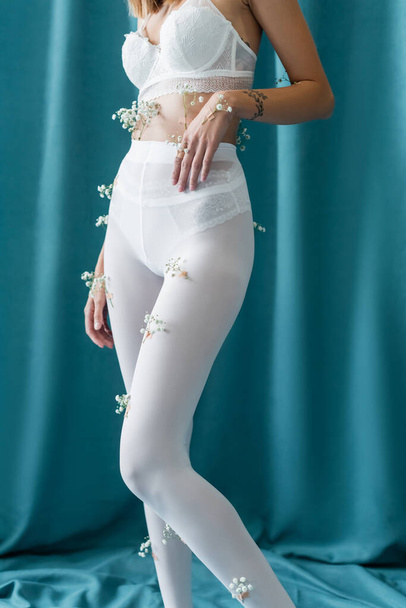 partial view of slim woman in lace bra and tights with white tiny flowers standing on green draped background - 写真・画像