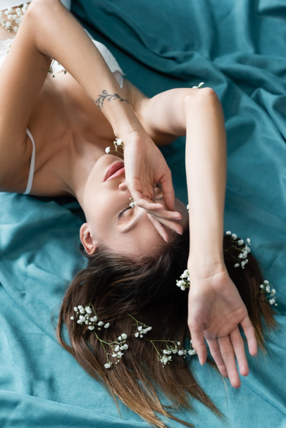 overhead view of woman with white gypsophila flowers on body and hair, lying with closed eyes on turquoise drape - Foto, afbeelding