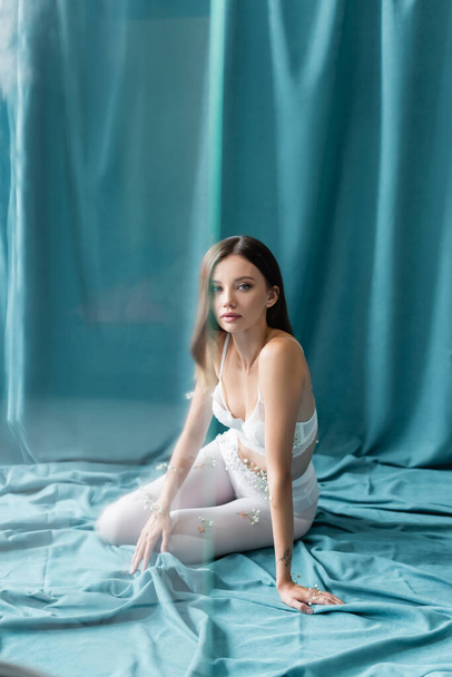 young woman in lace bra and white tights, with small flowers on body, looking at camera while sitting on green drapery - Photo, Image
