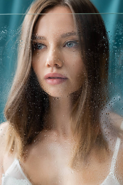close up view of young brunette woman posing near wet glass on turquoise background - Zdjęcie, obraz