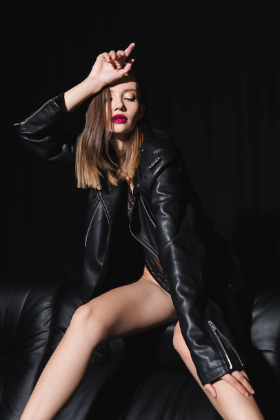 seductive woman in black bodysuit and leather jacket posing on couch on black background - Fotoğraf, Görsel