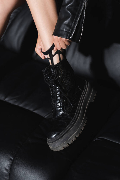 cropped view of woman in black leather boot adjusting sock near sofa - Photo, Image