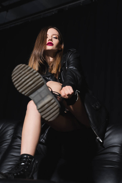low angle view of sexy woman adjusting boot while sitting on sofa on black background - Foto, Imagem