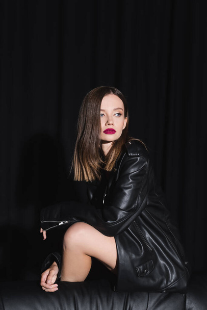 sensual brunette woman in leather jacket looking away while posing on black background with drape - Foto, afbeelding
