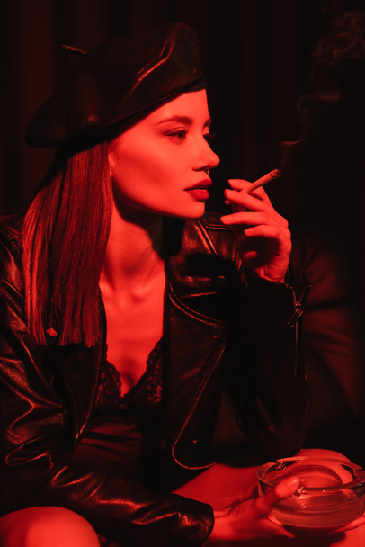 sexy woman in leather beret, jacket and bodysuit smoking in red light on black background - Zdjęcie, obraz