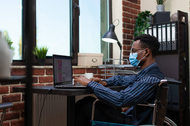 African american employee who is paralyzed wearing covid19 protection mask analyzing key performance indicators - Foto, immagini