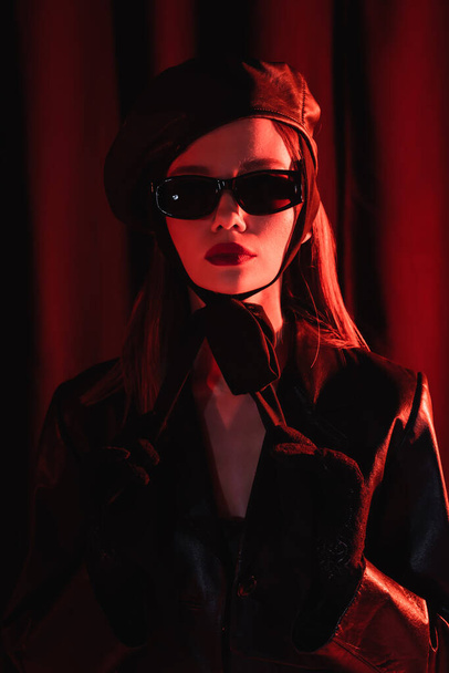 glamour woman in leather beret, black jacket, sunglasses and gloves posing on dark draped background - Photo, Image