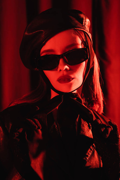 portrait of young woman in black sunglasses, leather beret and gloves on dark background with red light - Photo, Image