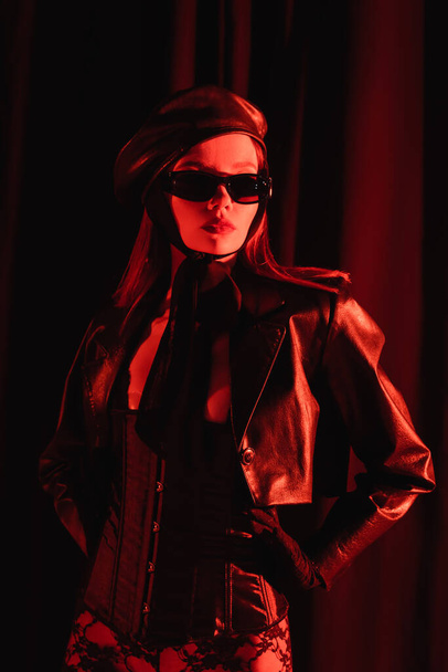 stylish woman in leather jacket, beret and black sunglasses standing with hands on hips on dark draped background with red light - Photo, Image