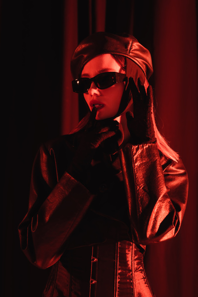 seductive and stylish woman in leather jacket, sunglasses and gloves touching lips on dark draped background - Фото, изображение