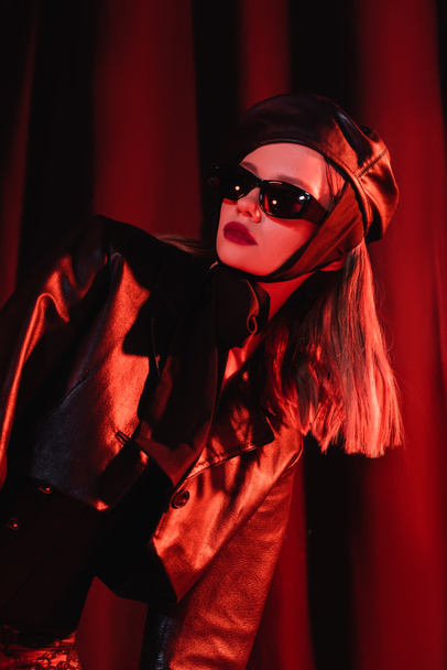 glamour woman in black sunglasses, leather jacket and beret posing on dark background with drapery and red light - Φωτογραφία, εικόνα